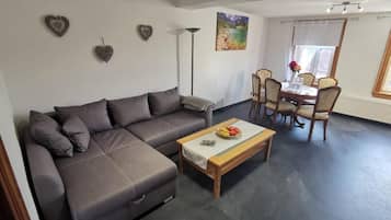 Ein modernes Wohnzimmer mit einem grauen Ecksofa, einem Couchtisch mit Obst und einem Essbereich mit Tisch und Stühlen.