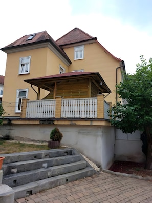Ein zweistöckiges Gebäude mit einem Balkon, einem kleinen Garten und einer Treppe aus Beton, die zum Eingang führt.