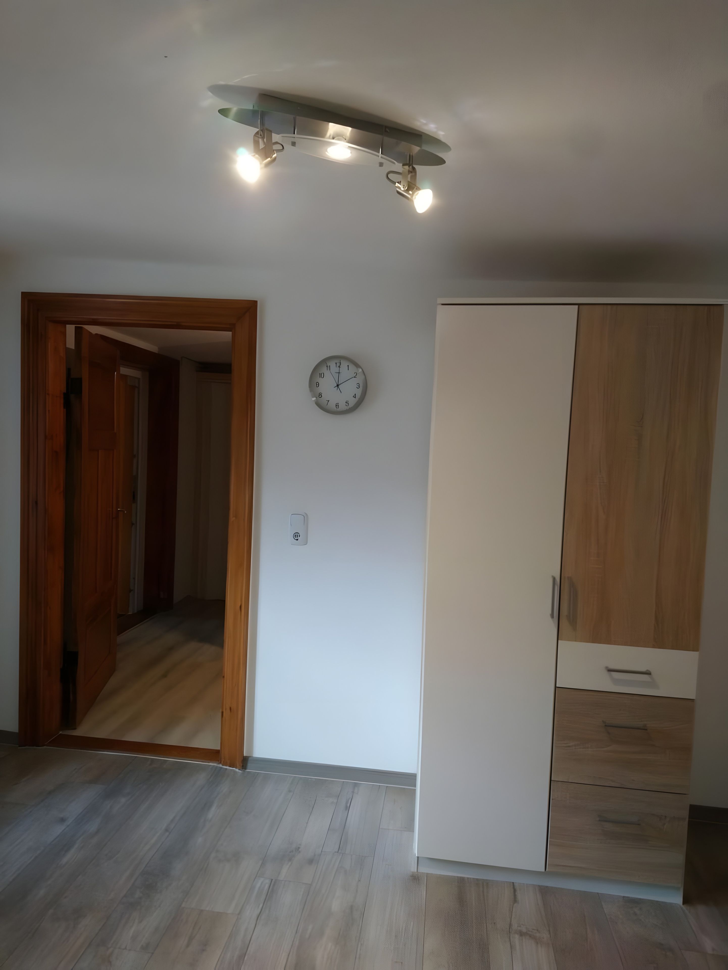 A room with a wooden wardrobe, a clock on the wall, and a ceiling light fixture.