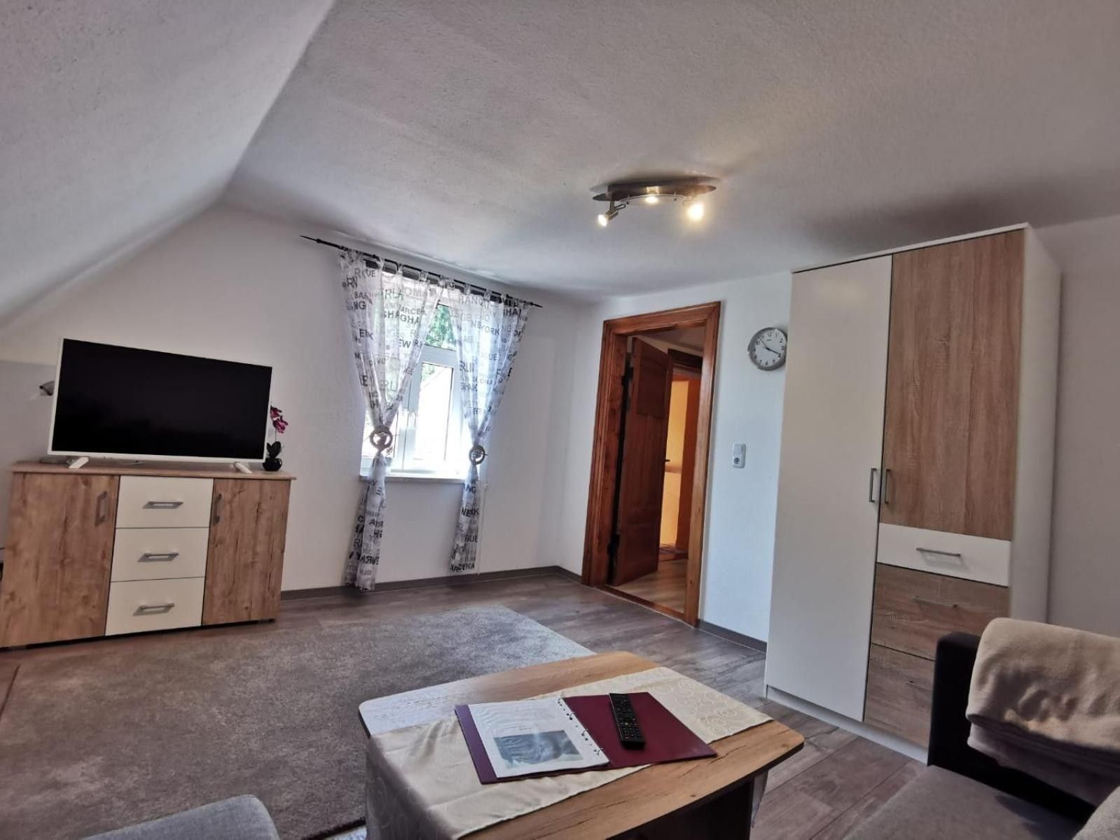 Ein Zimmer mit Fernseher auf einer Holzkommode, einem Schreibtisch mit Papieren und einer Fernbedienung, einem Kleiderschrank und einer Tür zu einem anderen Raum.