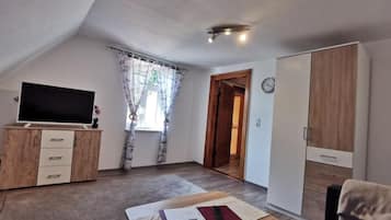 Ein Zimmer mit Fernseher auf einer Holzkommode, einem Schreibtisch mit Papieren und einer Fernbedienung, einem Kleiderschrank und einer Tür zu einem anderen Raum.