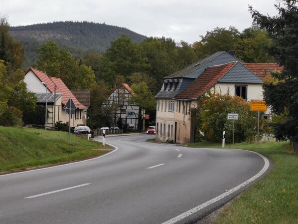 A rural road curving through a village with traditional houses, a clear sky, and a forested hill in the background.