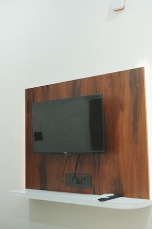 A flat-screen TV mounted on a wooden panel wall with a white shelf below. - Hotel SP Residency (Indore)