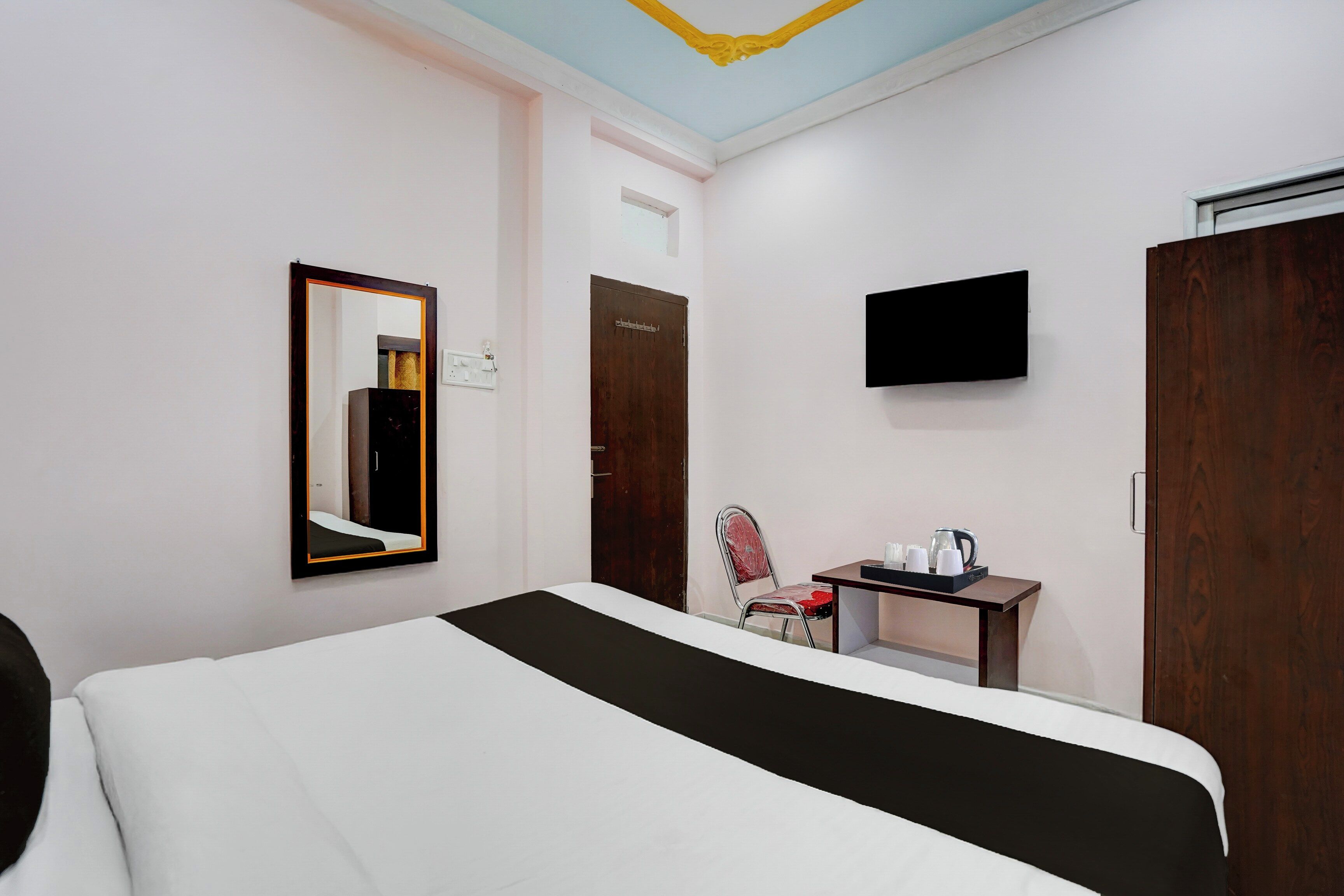 A hotel room with a bed, a chair, a table with a tray of cups, a television, and a wardrobe.