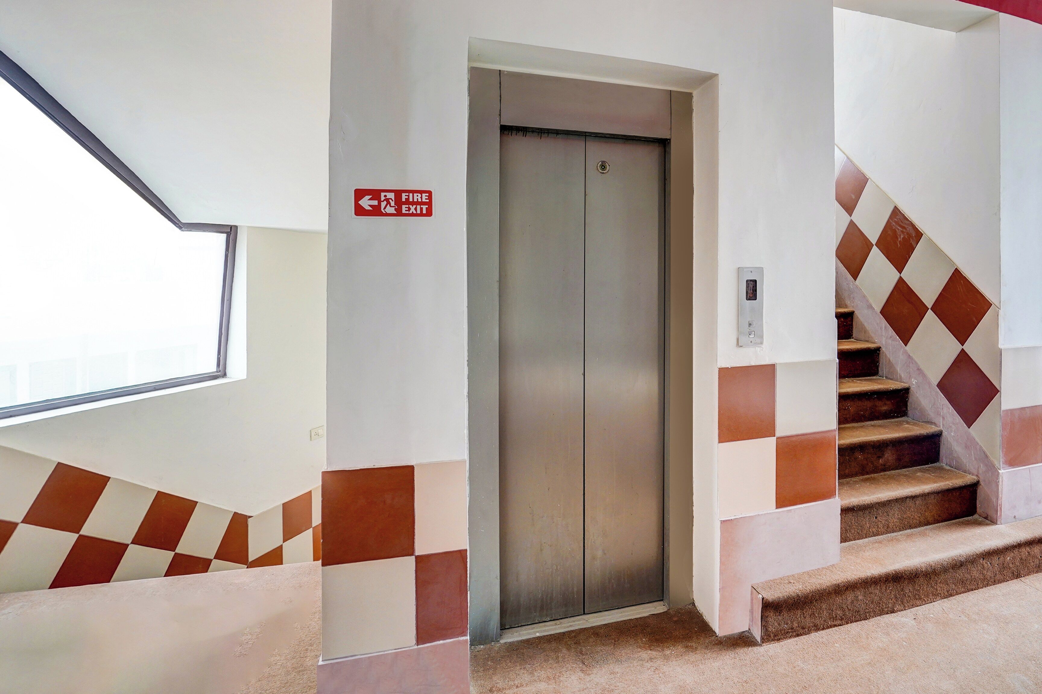 An elevator with a fire exit sign, a window, and a staircase with patterned tiles.