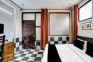 A hotel room with a checkered floor, a bed with black and white bedding, a wooden dresser, a television, and a window with blinds and curtains. - Hotel O Sector 93 Noida (Noida)
