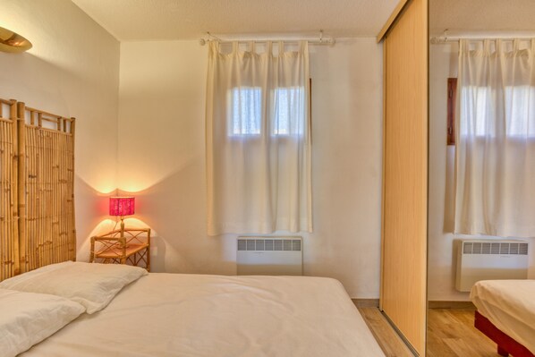 A hotel room with a bed, a bedside lamp, a window with curtains, an air conditioning unit, and a mirror. - By the Water...!!! 50 Meters from the Beach (Narbonne-Plage)