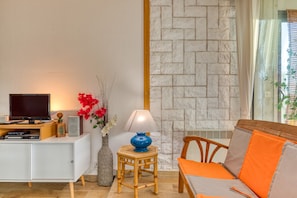 Una sala de estar moderna con un sofá blanco, un televisor sobre un mueble blanco, una lámpara azul sobre una mesita de madera y un jarrón decorativo con flores.