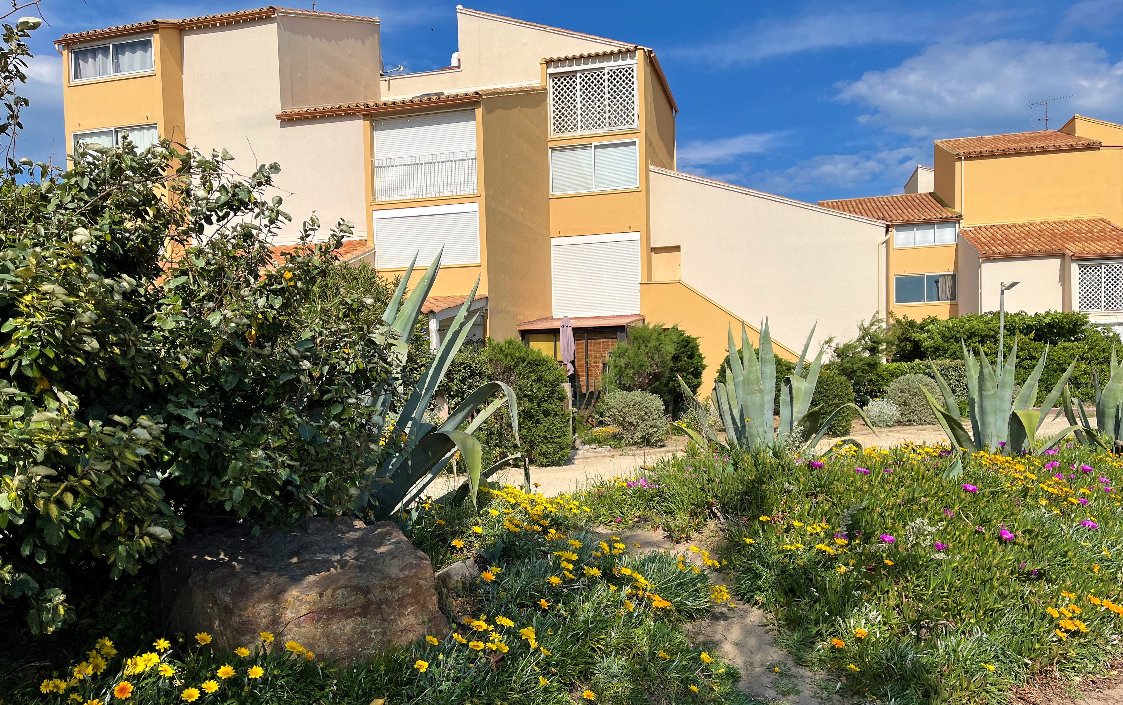 Un immeuble à plusieurs étages doté de balcons, entouré d’un jardin où poussent différentes plantes et fleurs.