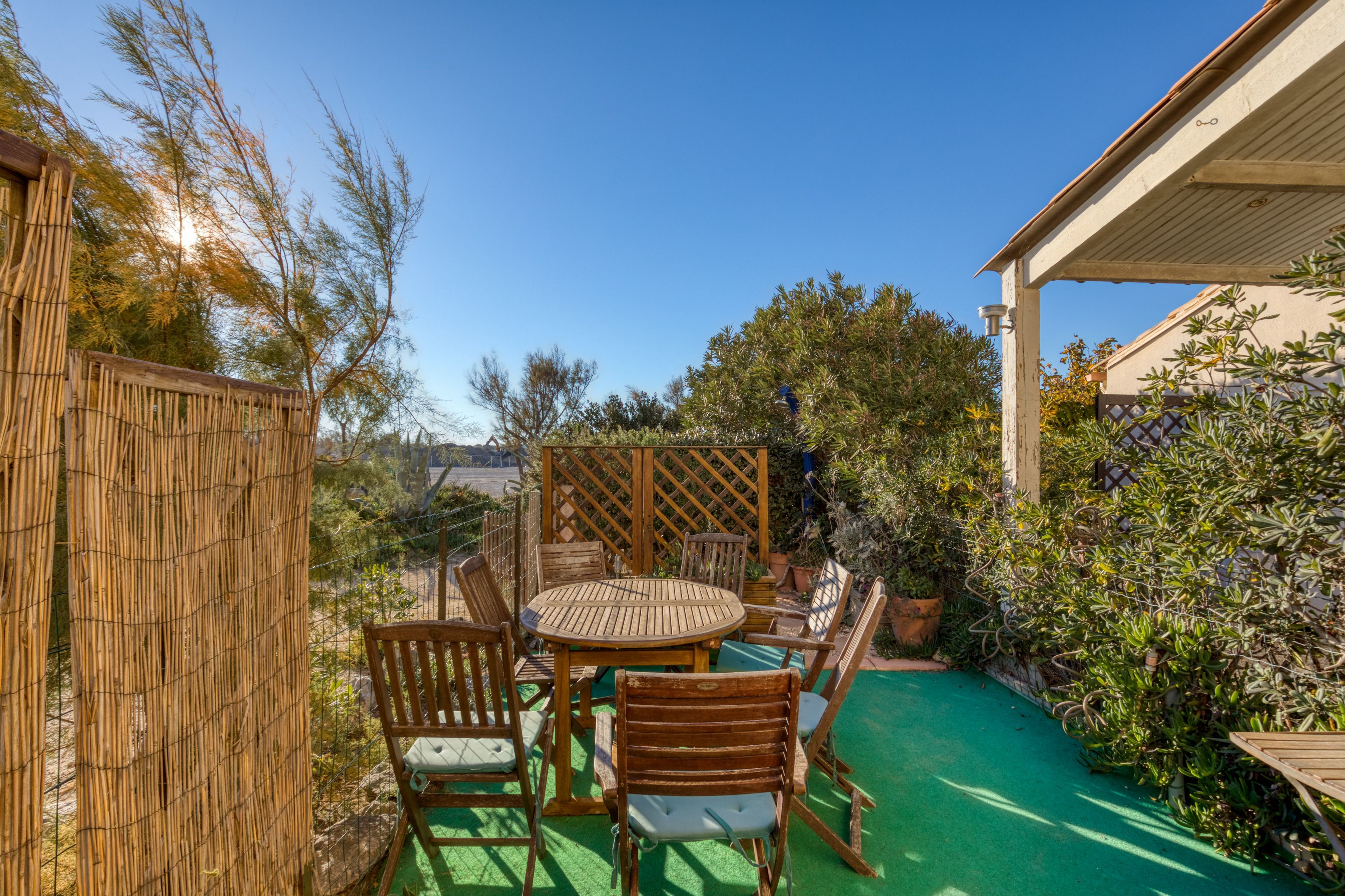 A patio with a round wooden table and chairs, surrounded by a bamboo fence and various plants.