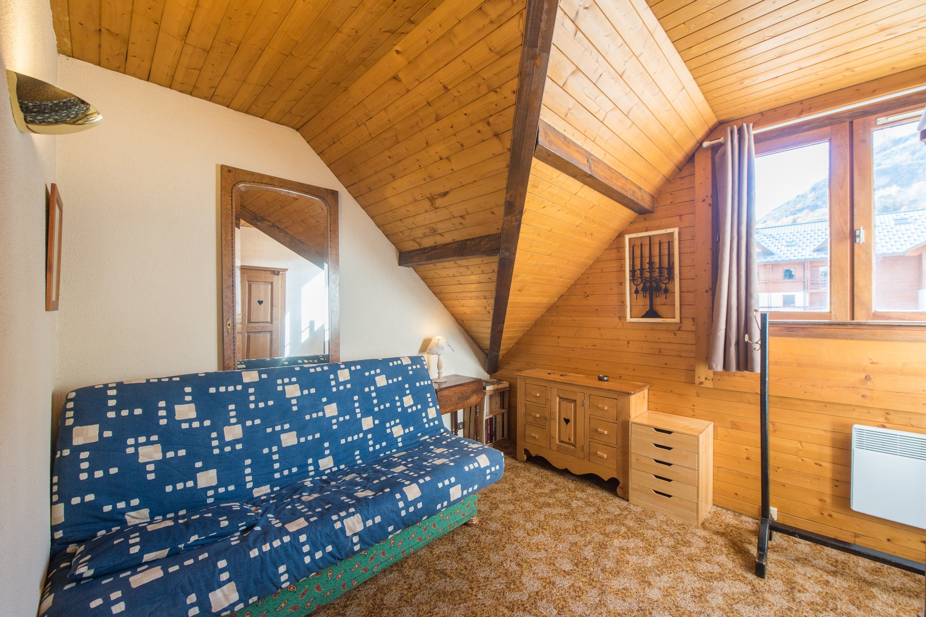 A cozy room with a sloped wooden ceiling, a blue patterned sofa, a wooden cabinet, and a window with curtains.
