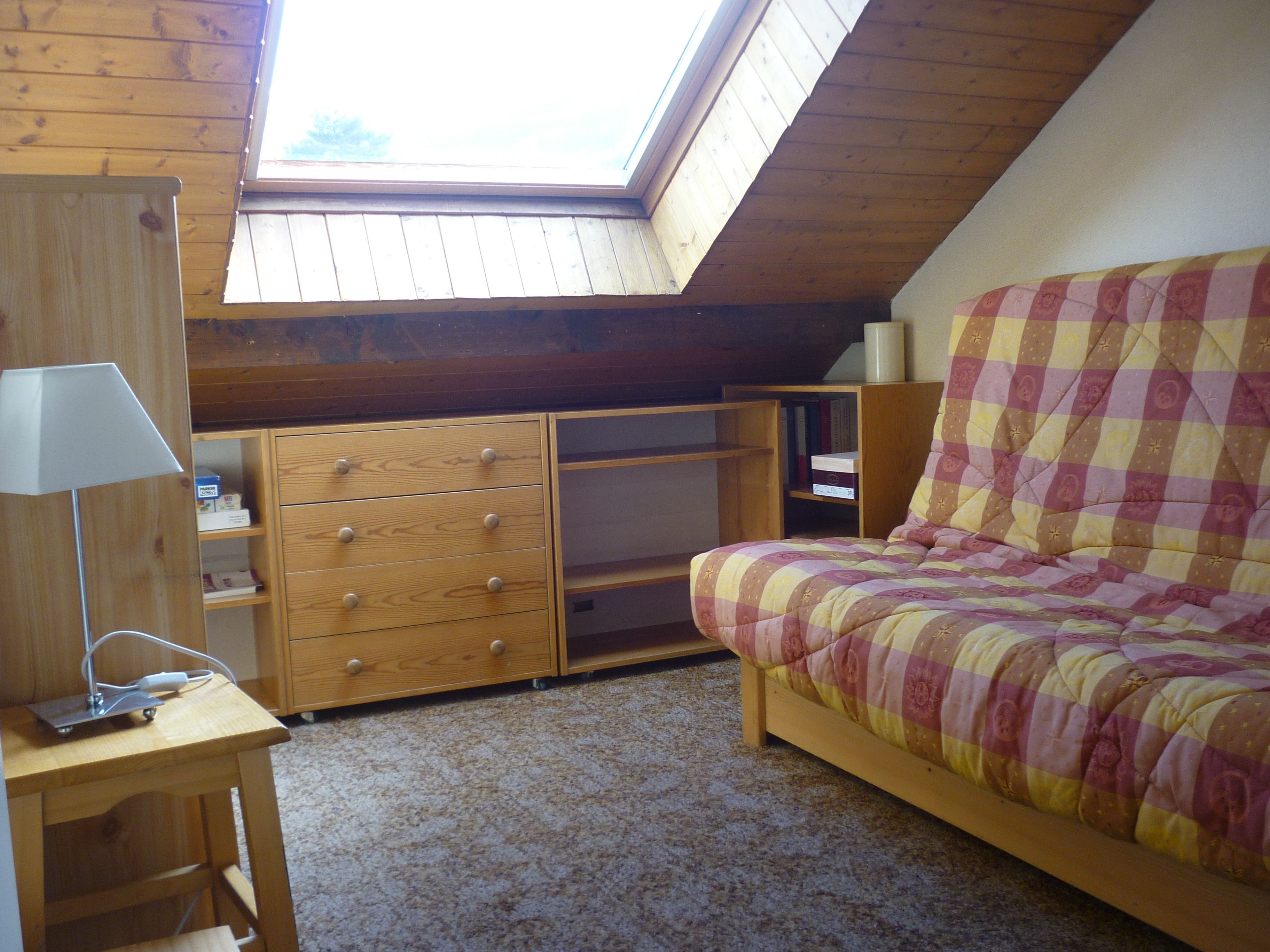 A bedroom with a sloped ceiling, a bed with a checkered bedspread, a wooden dresser with drawers, and a desk with a lamp.