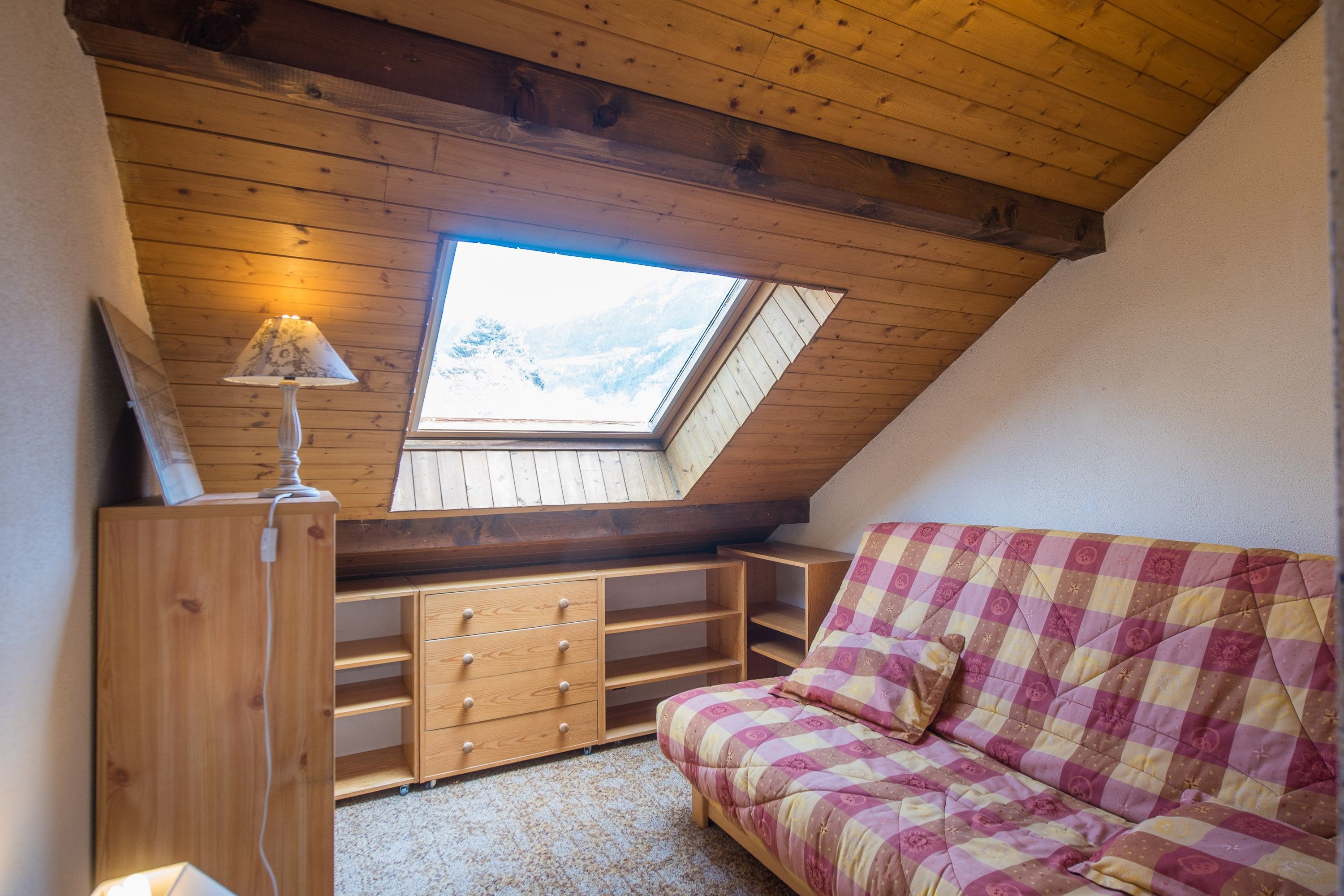 A cozy room with a sloped ceiling, a skylight, a wooden dresser, a sofa, and a lamp.