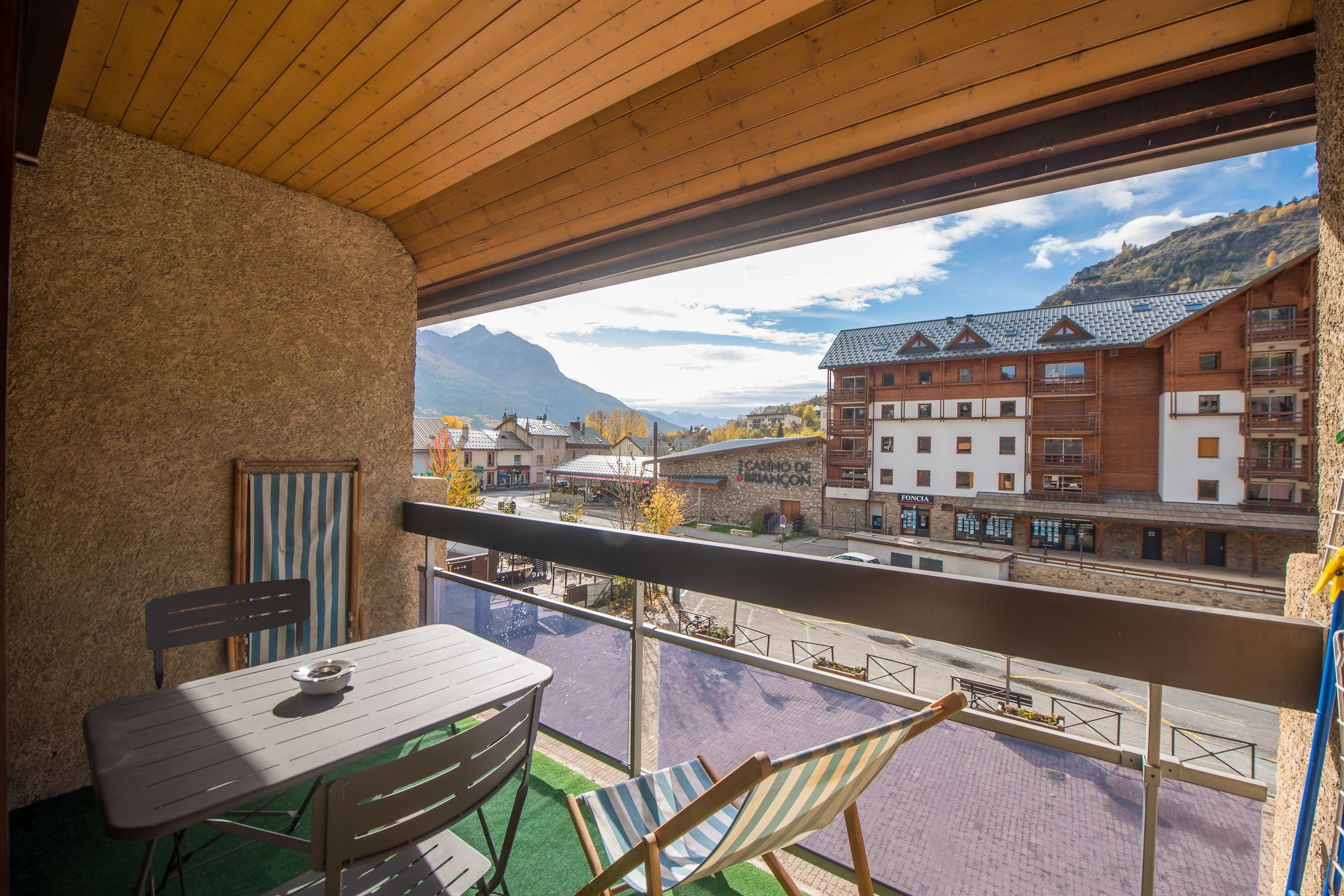 Un balcó amb taula i cadires, vista a una vila amb edificis i muntanyes, i un sostre de fusta.