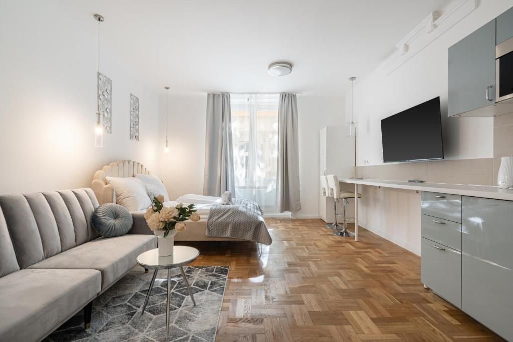 Een moderne hotelkamer met een bank, een bed, een kleine tafel met bloemen, een kitchenette en een televisie.