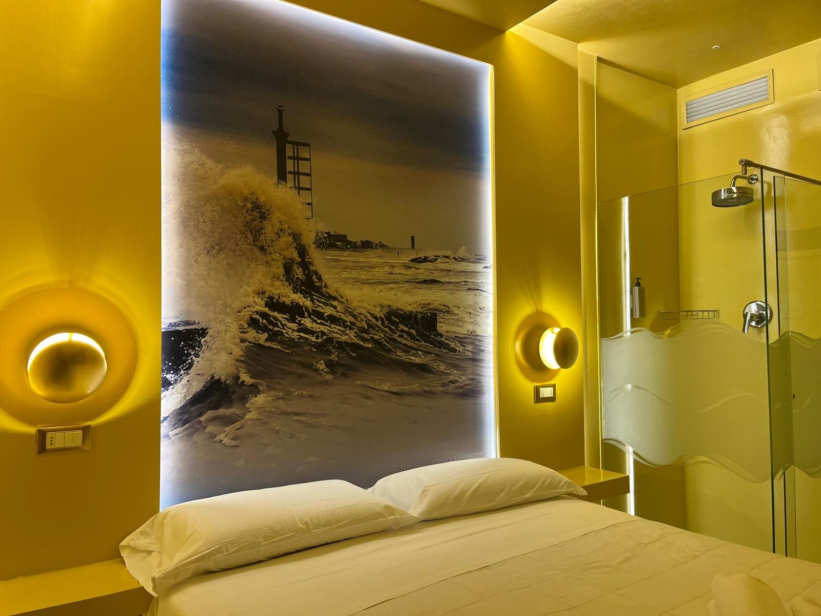 Una camera d'albergo con un letto, un grande affresco raffigurante un faro e onde, e un box doccia in vetro.