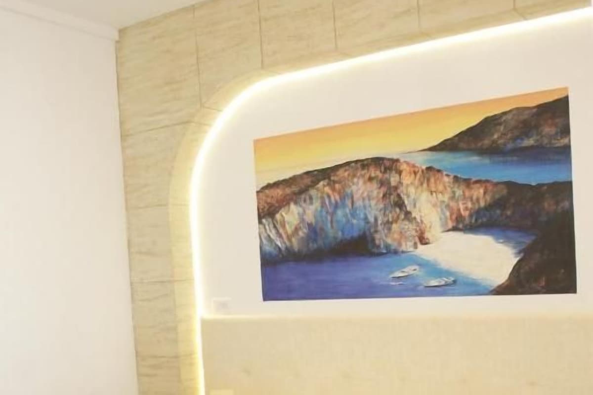 Una cama con colcha blanca acolchada, una mesita de noche con lámpara y un cuadro de un paisaje costero en la pared.