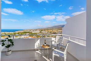 A balcony with a view of a coastal city, a table with a bottle and glass, and white chairs. - Island Village Paradise apartment (Costa Adeje)