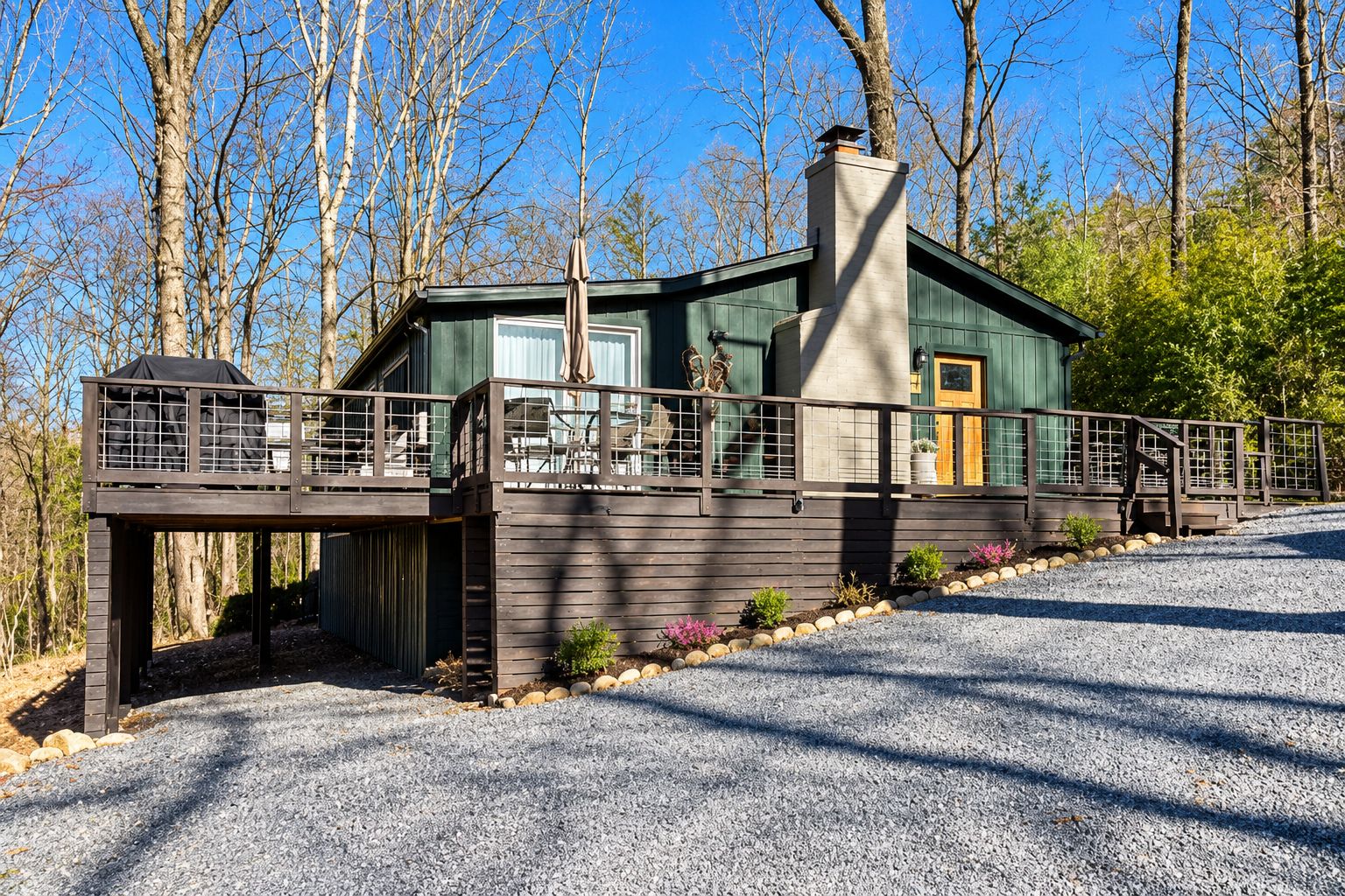 A cabin with a deck, a chimney, and a gravel driveway.