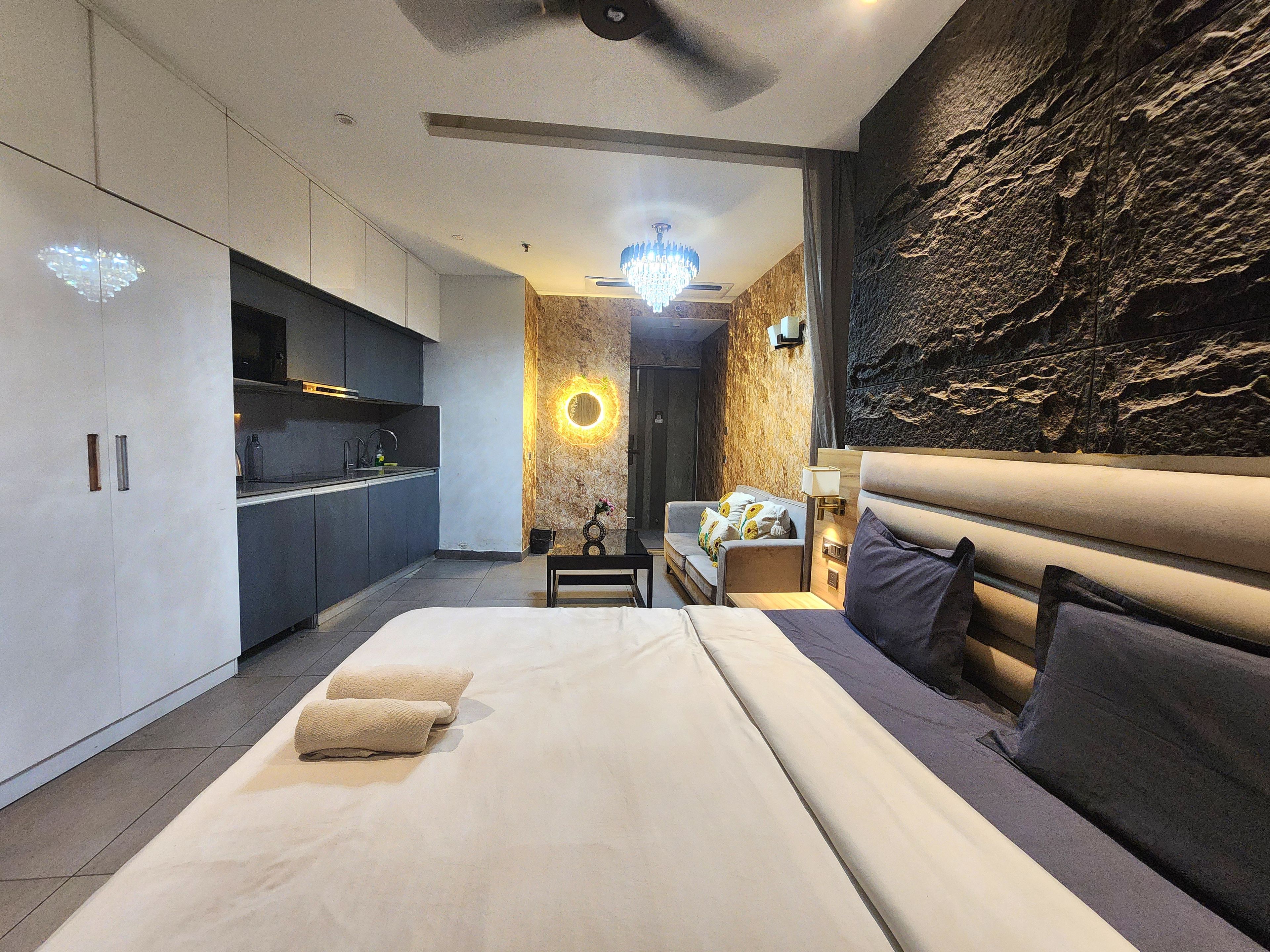A modern hotel room with a large bed, a sitting area with a sofa, a small table, and a kitchen area with cabinets and a microwave.