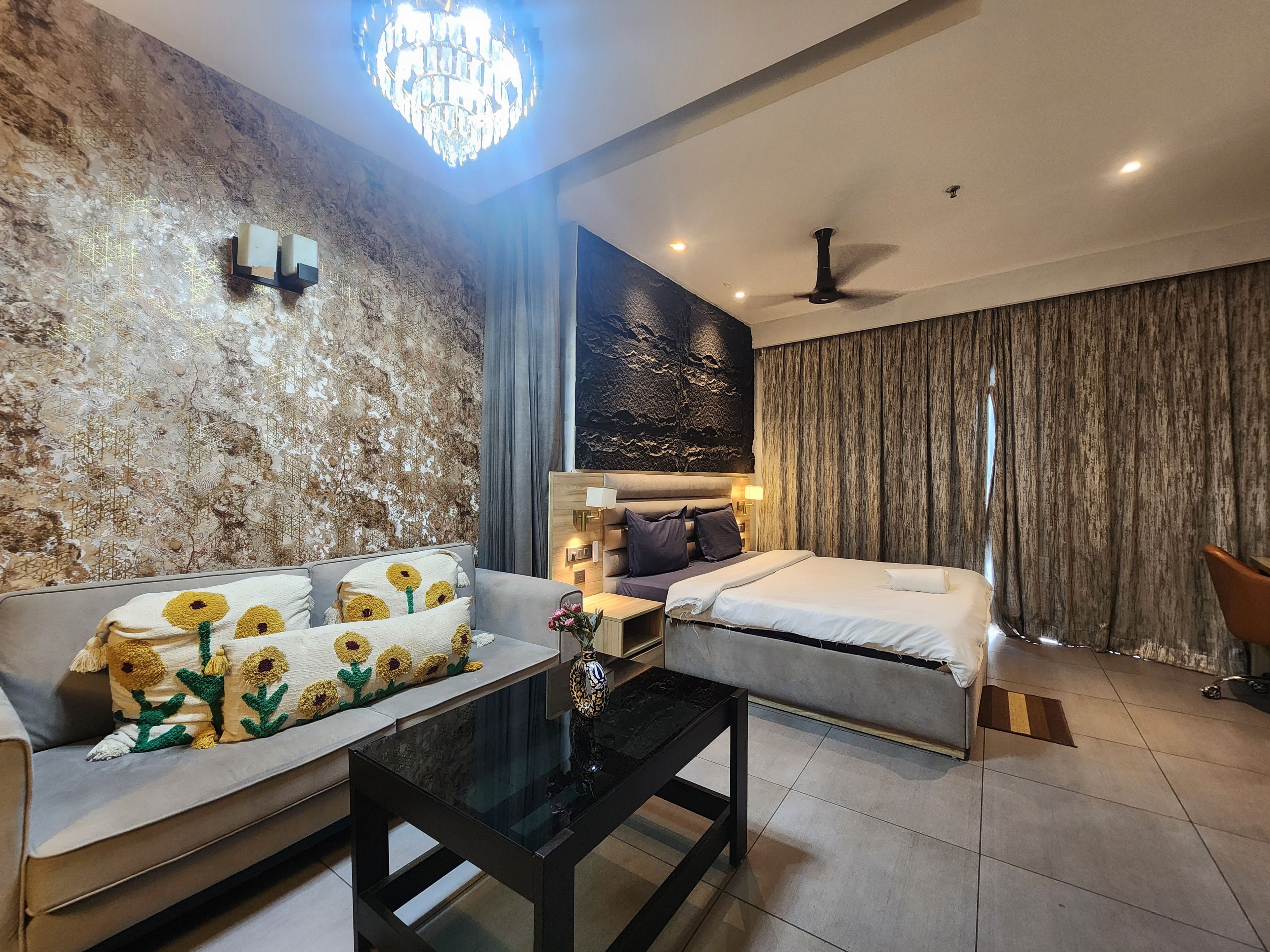 A modern hotel room with a sofa, a bed, a glass coffee table, and a chandelier.