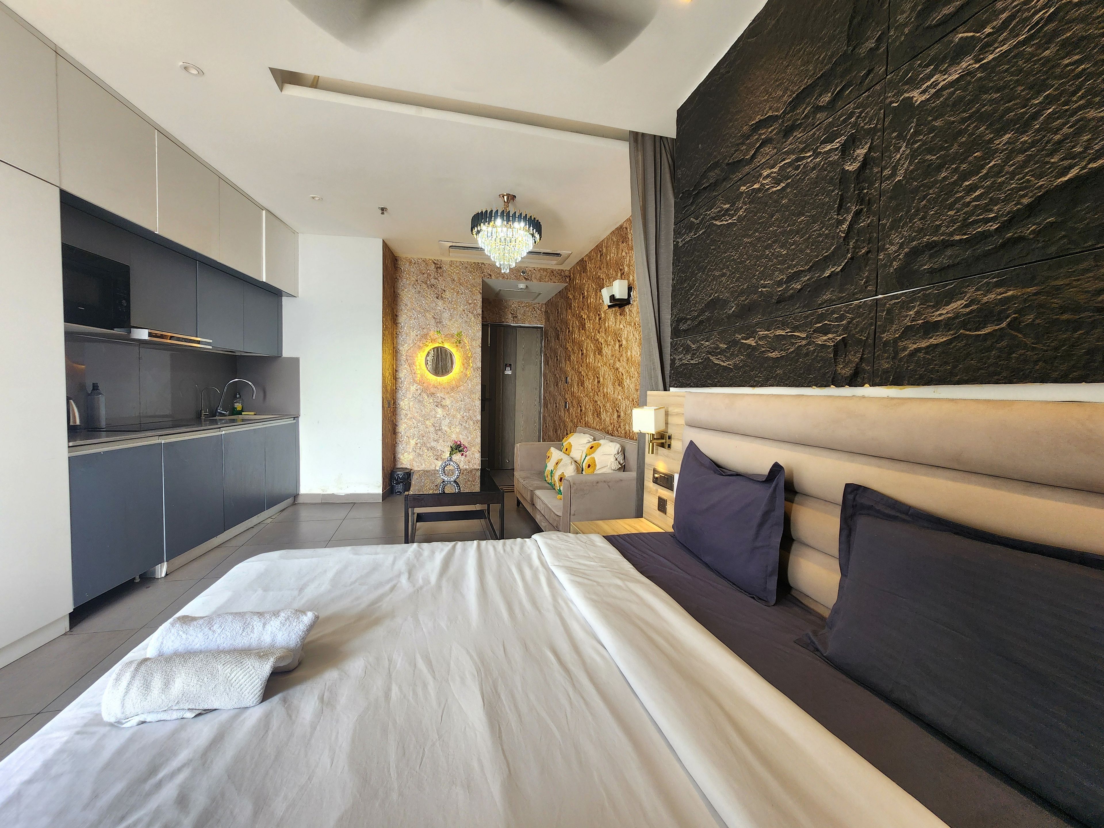 Habitación de hotel moderna con una cama grande, una kitchenette independiente, un muro decorativo y una lámpara en el techo.
