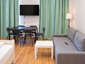 Ein Zimmer mit einem wandmontierten Flachbildfernseher, einem Esstisch mit Stühlen, einem Sofa, einem weißen Couchtisch und einer Lampe.