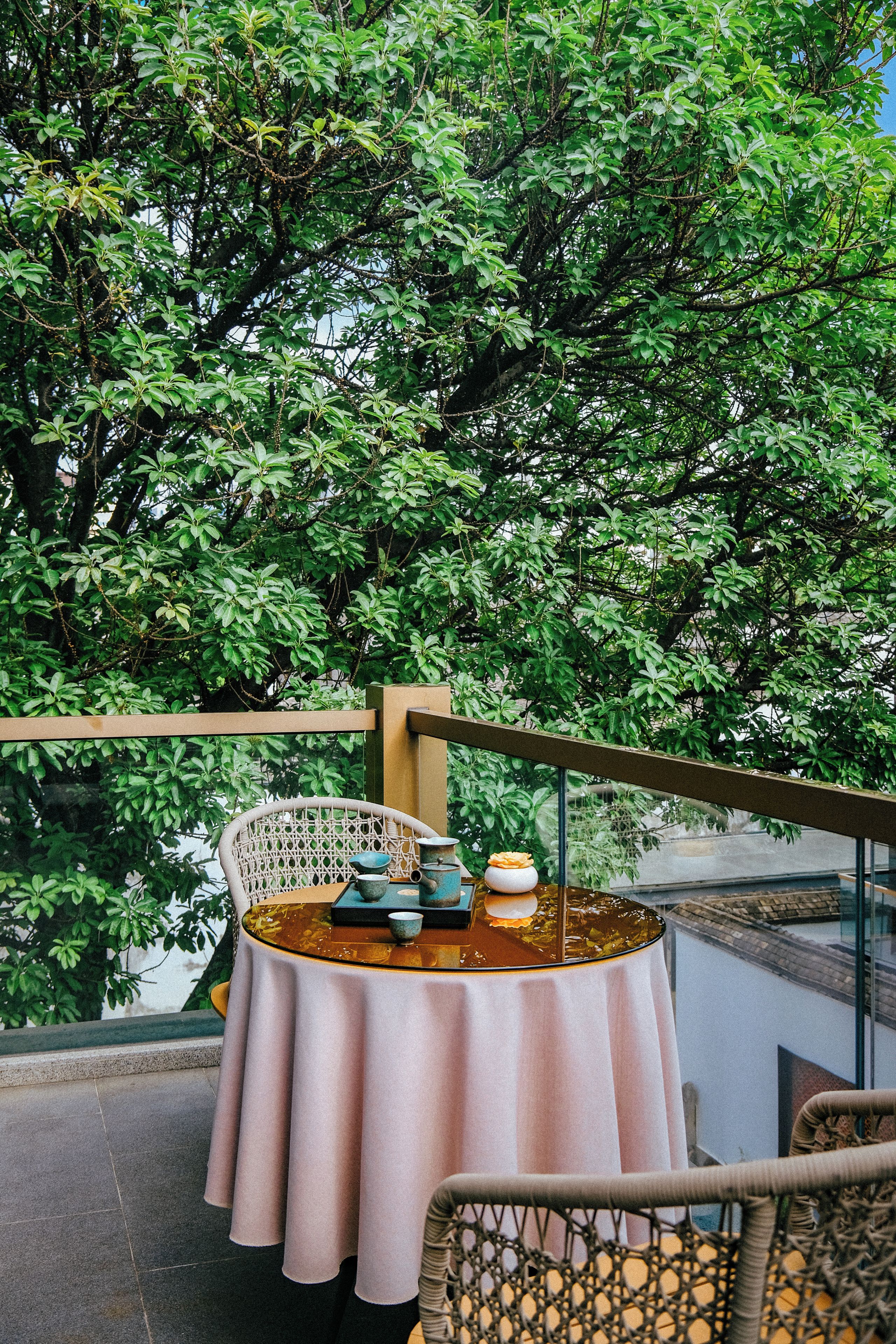 A round table with a white tablecloth, a wicker chair, and a view of a lush green tree.