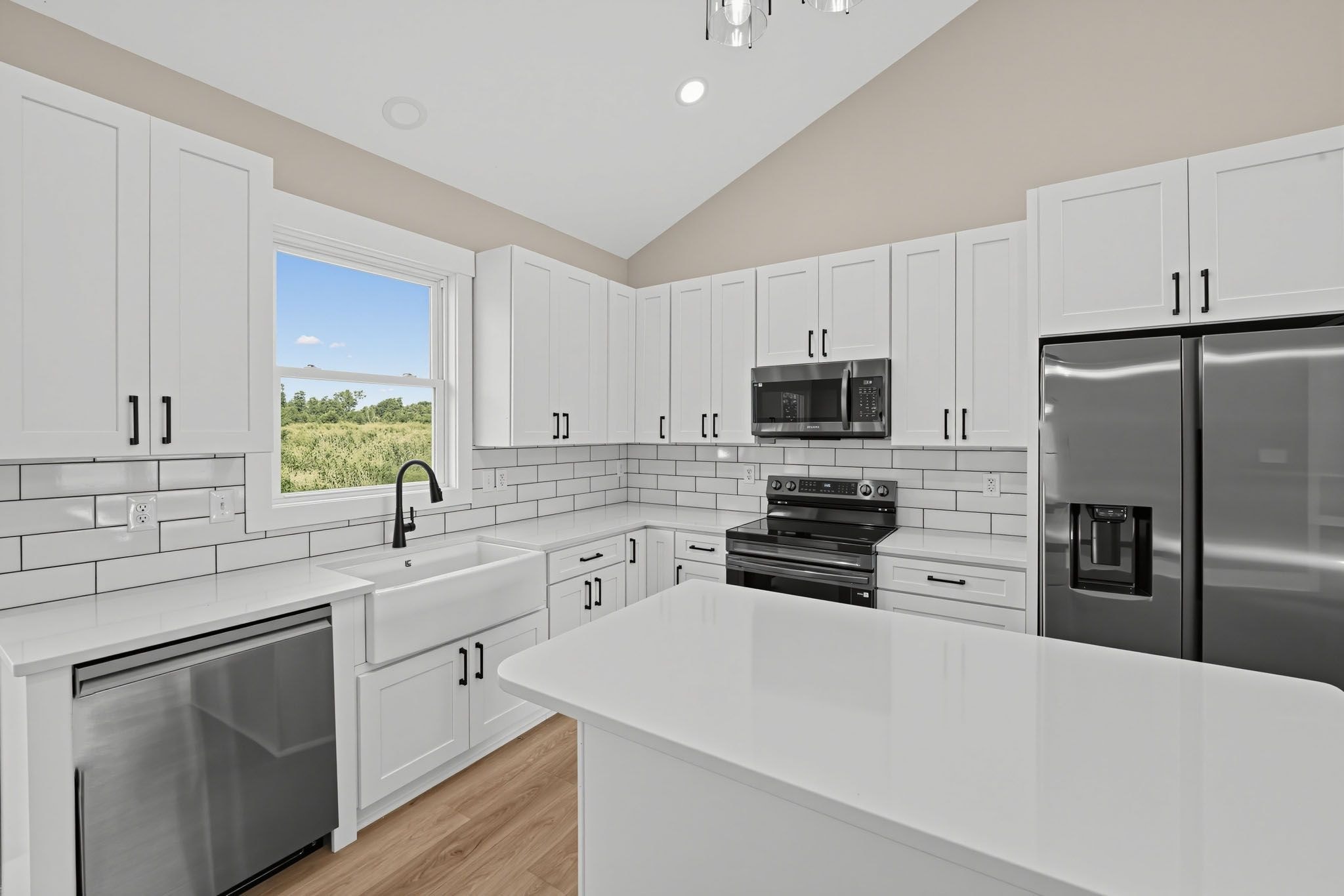 A modern kitchen with white cabinetry, a stainless steel refrigerator, a built-in oven, and a window with a view of greenery.