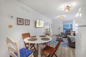 A well-lit living area with a dining table, chairs, a sofa, and a television mounted on the wall. - Gulf Shores Cozy Getaway: 1BR Condo Pool Access/5 Minute Walk to Beach (Gulf Shores)