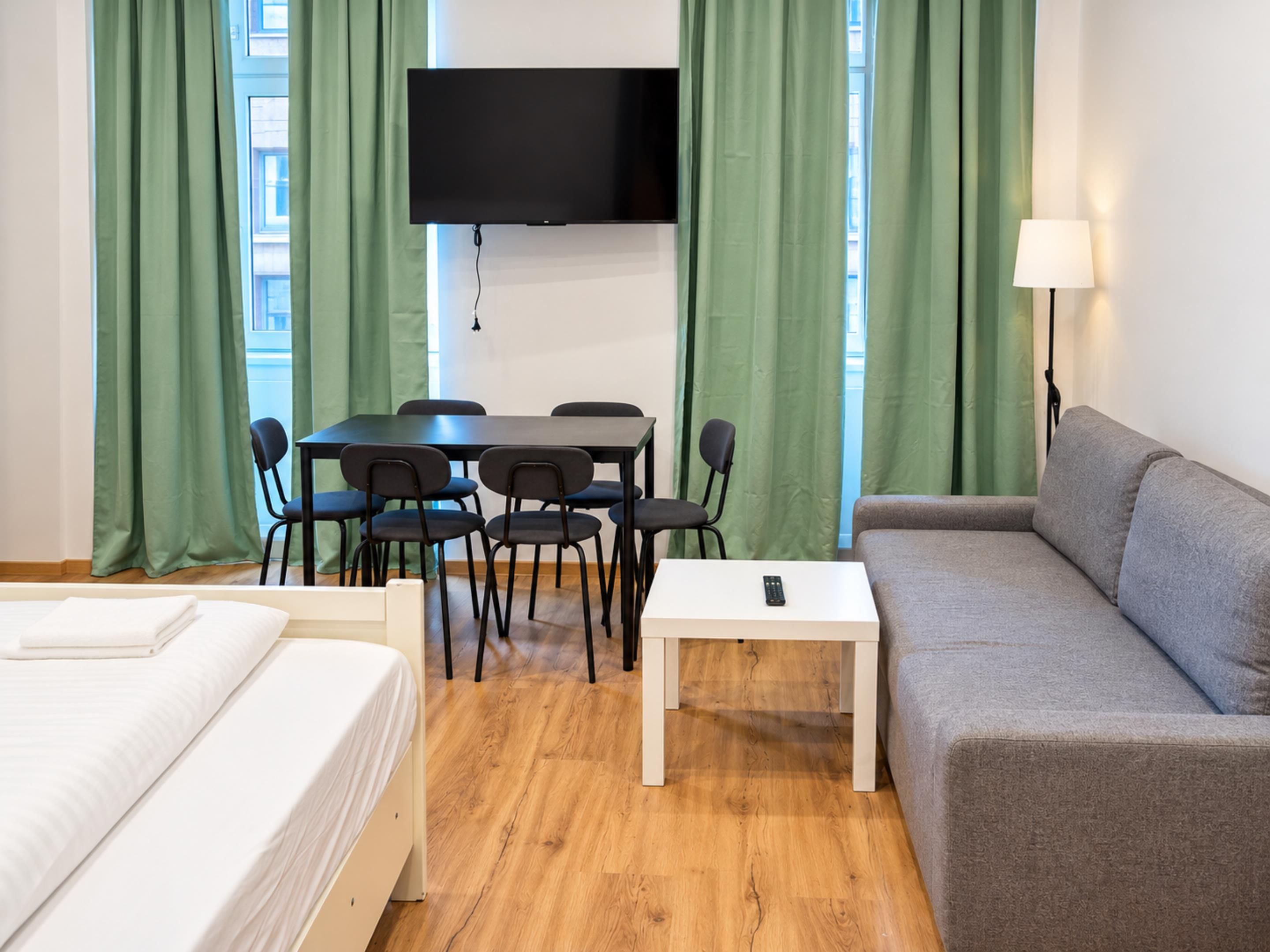 A hotel room with a bed, a sofa, a dining table with chairs, a TV mounted on the wall, and a lamp.