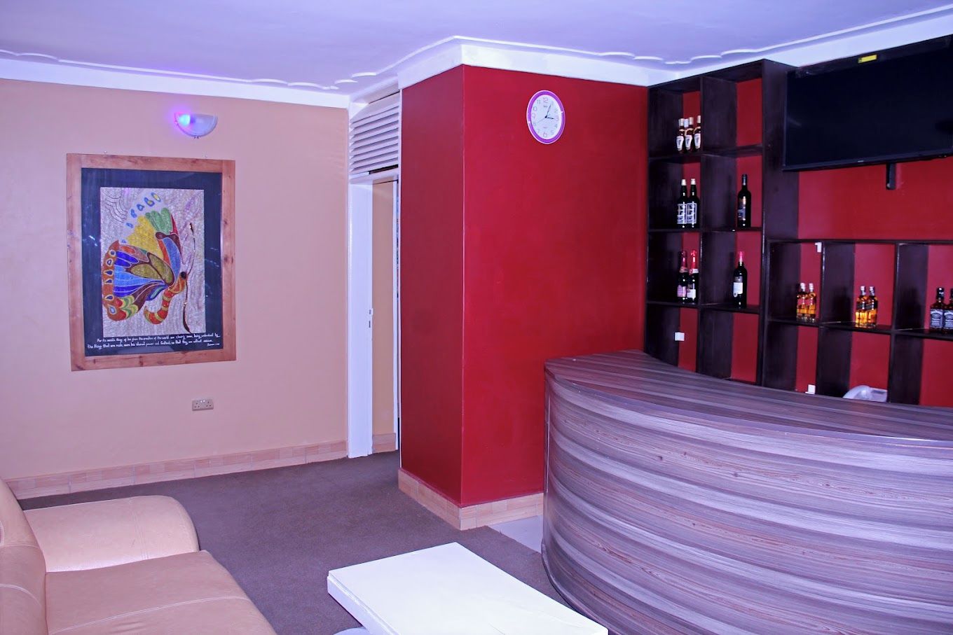 A room with a red wall, a clock, a bar area with bottles, a sofa, and a framed artwork.