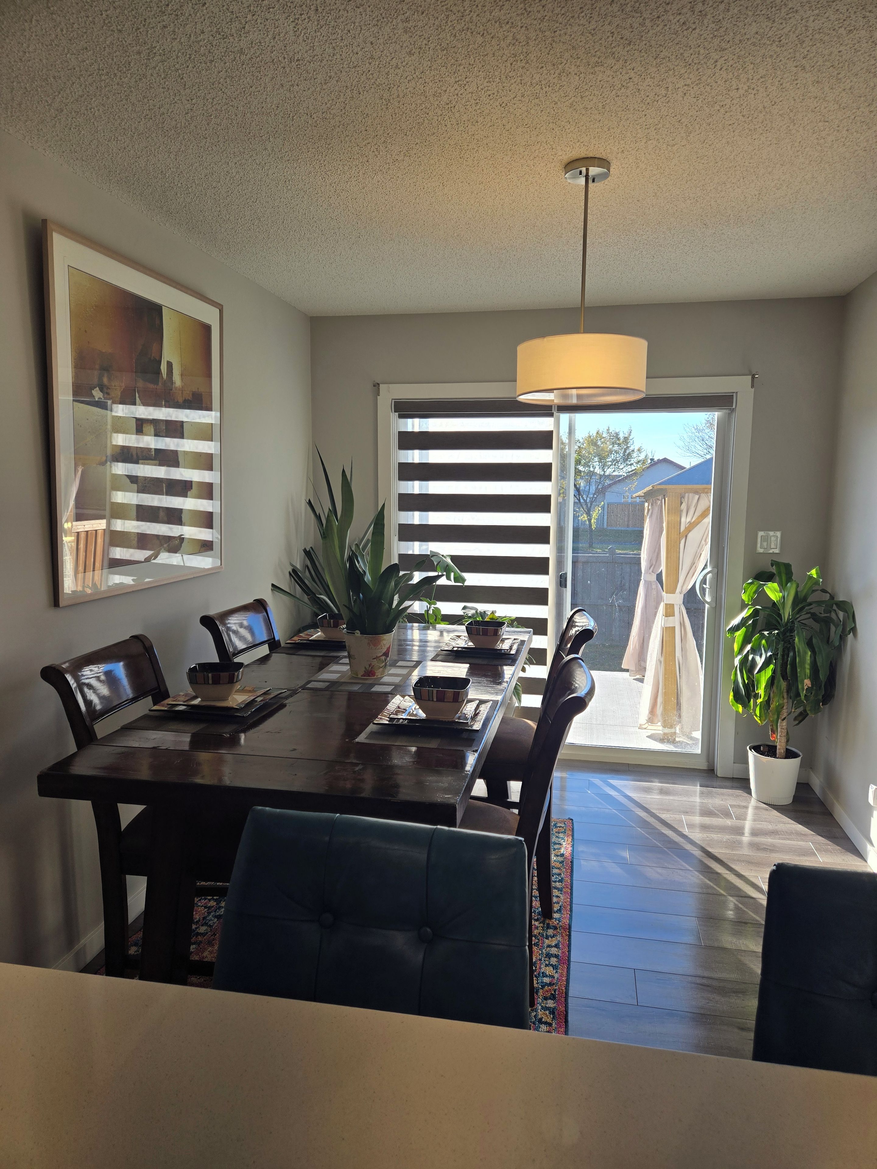 A dining area with a table, chairs, a hanging light fixture, and a view through a window with blinds.