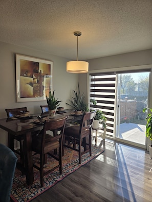 A dining area with a wooden table, chairs, a hanging light fixture, and a large window with blinds. - Modern home w/ Full Kitchen & In-Unit Laundry (Edmonton)