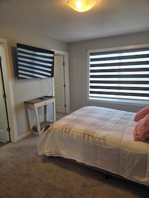 A bedroom with a bed, a desk, a TV, and a window with blinds. - Modern home w/ Full Kitchen & In-Unit Laundry (Edmonton)