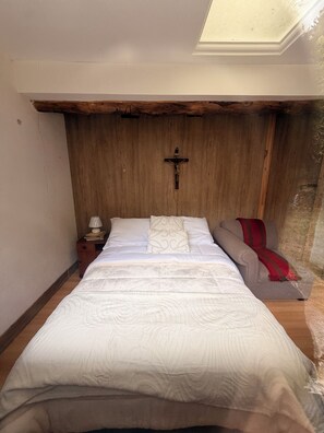 A bedroom with a wooden ceiling, a cross on the wall, a bed with white bedding, a nightstand with a lamp, and a red throw blanket on a couch. - Hacienda Molobog: 3BR near Cuenca, Mountain Views & Gardens (Azogues)