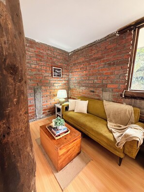 A room with a brick wall, a green sofa, a wooden coffee table with books and a plant, and a window with a view. - Hacienda Molobog: 3BR near Cuenca, Mountain Views & Gardens (Azogues)