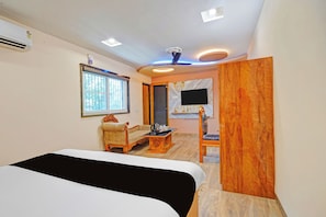 A hotel room with a bed, a sofa, a wooden chair, a television mounted on the wall, and a ceiling fan.