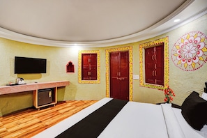 A hotel room with a bed, a desk with a flat-screen TV, a refrigerator, and a wall with a decorative pattern.