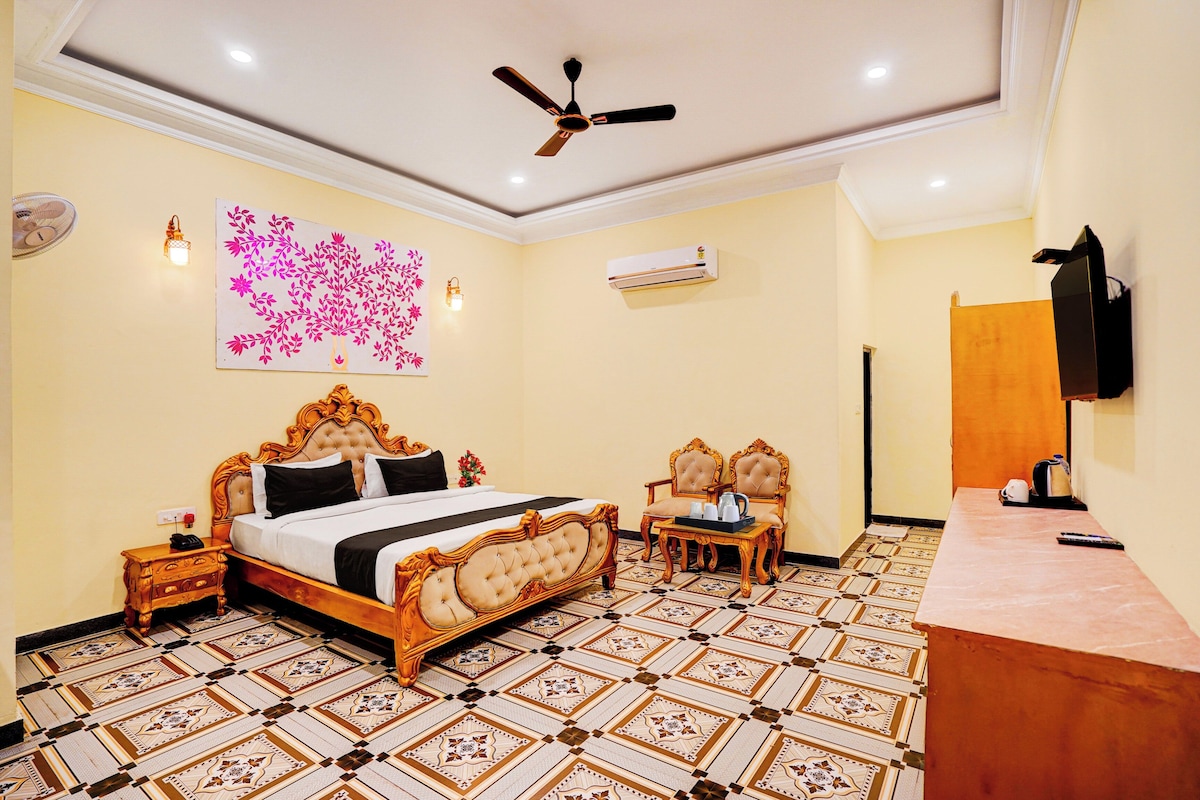 A hotel room with a large bed, a desk, two chairs, a ceiling fan, an air conditioning unit, and a decorative wall piece.