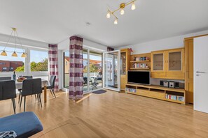 Ein modernes Wohnzimmer mit Holzboden, einer Essecke mit Tisch und Stühlen, einer TV-Konsole mit Büchern und Elektronik sowie einem großen Fenster mit Vorhängen.