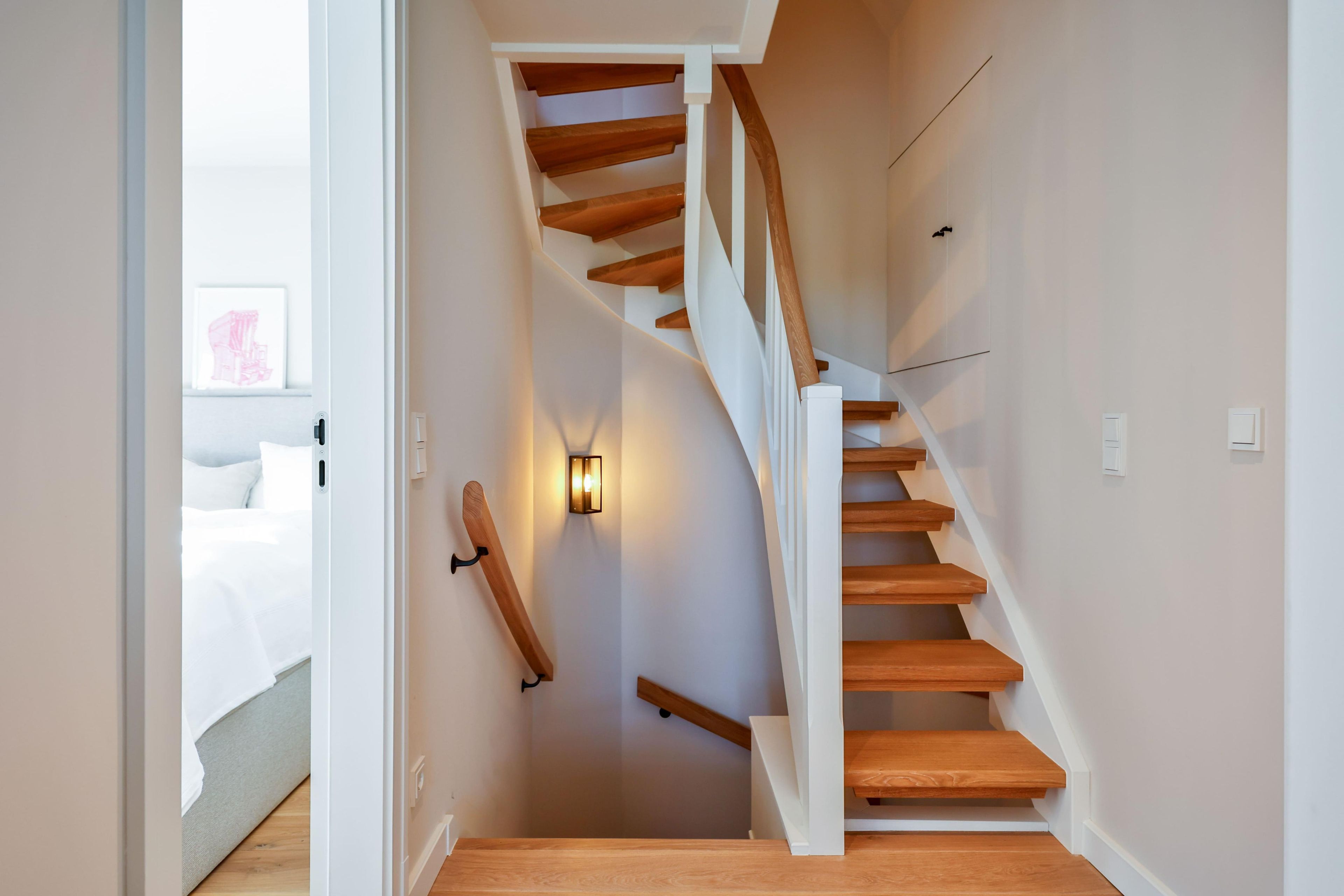 A staircase with wooden steps and white walls, leading to a bedroom with a bed and a wall-mounted light fixture.