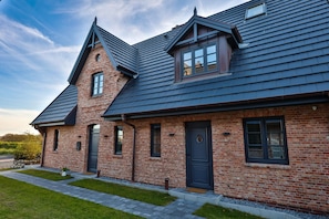 A two-story brick house with a dark shingle roof, a front door, and a small porch. - Stylish living with a view of the salt marshes, including a sauna, fireplace, and terrace (Sylt)
