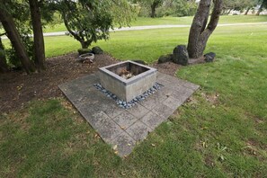 A square stone fire pit with a metal frame, surrounded by a stone border and small rocks, set on a paved area with grass around. - Wooded golf Course Retreat (Westerville)