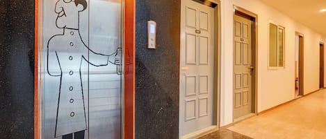 A hotel hallway with a glass elevator door featuring a simple line drawing of a man holding a drink.
