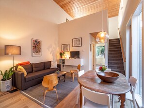 A modern living room with a sofa, coffee table, and dining table. There is a TV on a stand, a plant, and artwork on the walls. A staircase is visible in the background. - Mt. Tabor Guesthouse | Courtyard & Park Walk (Portland)