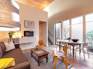 A modern living room with a flat-screen TV, a sofa, a coffee table, and a dining area with a table and chairs. - Mt. Tabor Guesthouse | Courtyard & Park Walk (Portland)