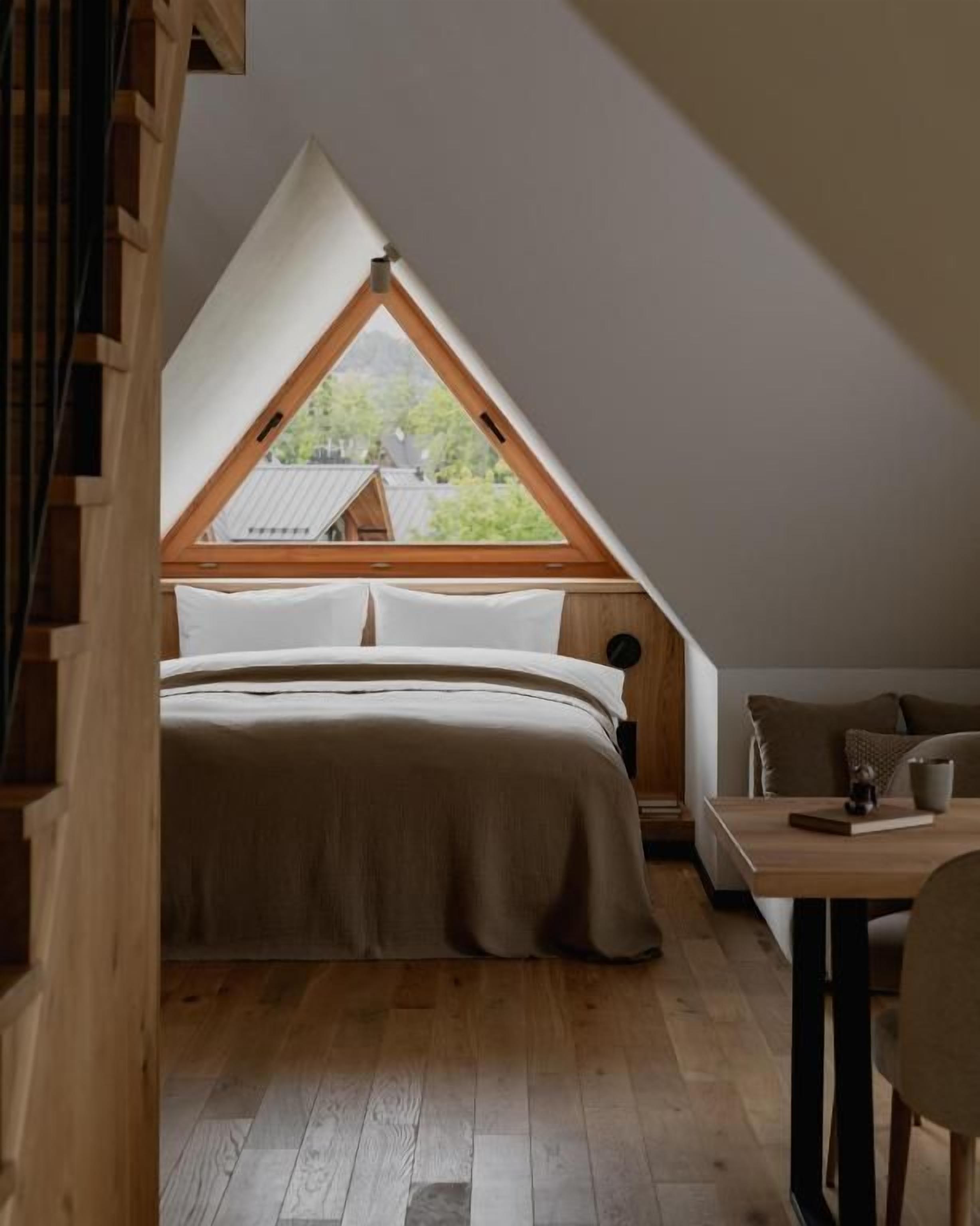 Ein Schlafzimmer mit schrägem Dach und einem großen Fenster, ein Bett mit weißer Bettwäsche sowie ein Holztisch mit einem Stuhl.
