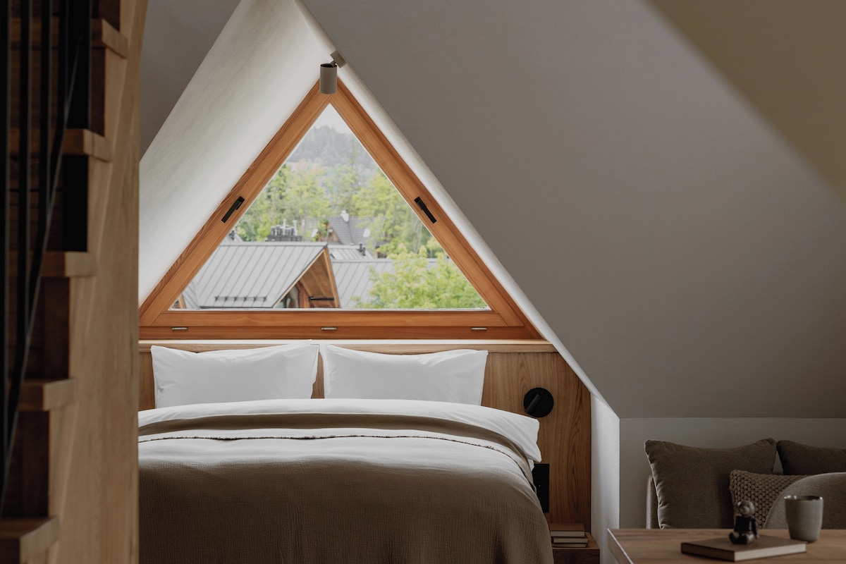 Quarto com teto inclinado, uma cama, uma mesa e vista para o exterior.