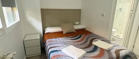 A single bed with a patterned bedspread, a nightstand, and a window with blinds.